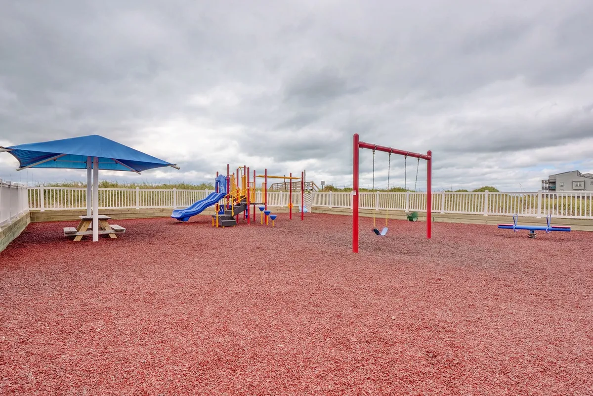 This image features a playful playground complete with swings, vibrant equipment, a picnic table, and a shade structure. Children are seen enjoying the area, with families gathered for picnics and social interactions under sunny skies.