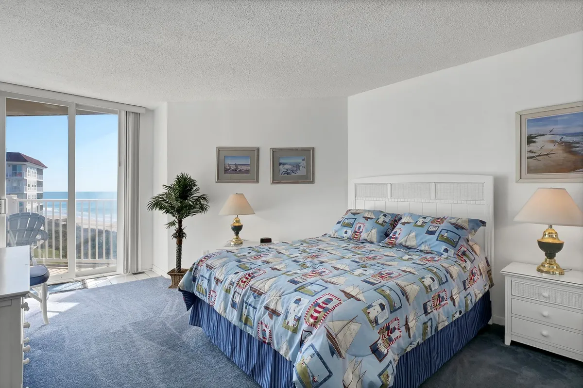 This image depicts a serene bedroom with nautical-themed bedding, a large window offering an ocean view, a potted plant, and a nightstand with a lamp. Sunlight floods the room, creating a tranquil and inviting atmosphere. The decor is distinctly coastal, aligning with the theme of restful seaside retreats.