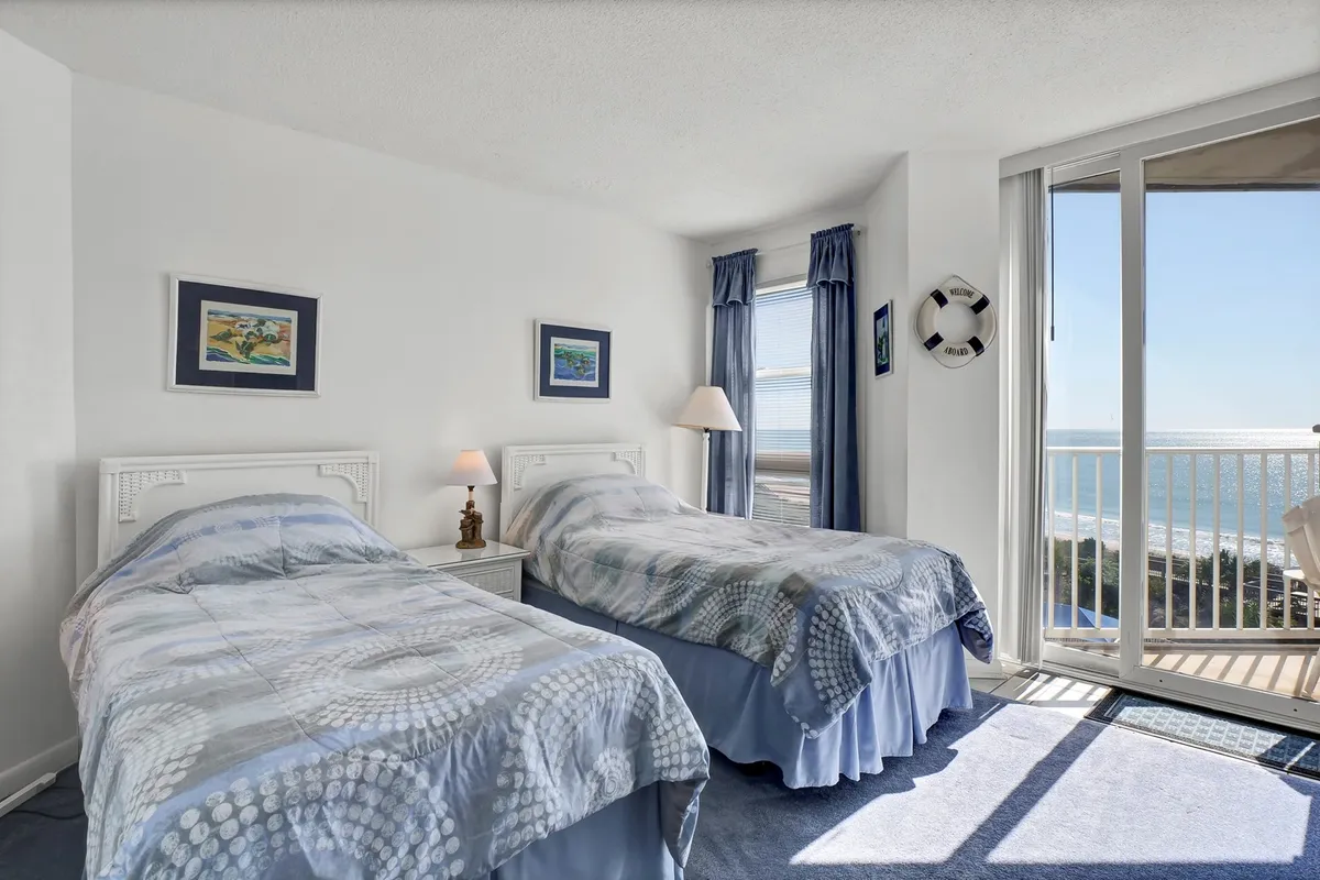 This image shows a guest bedroom with two twin beds, cheerful coastal artwork, a large window with an ocean view, and a lamp. The overall mood is calm and peaceful, making it an ideal space for children or friends.
