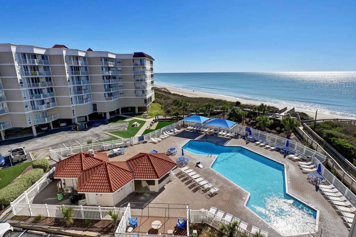 The image showcases a sparkling saltwater pool surrounded by inviting lounge chairs, with the condo building rising just steps from the sandy beach. The ocean stretches beyond, bathed in the warm glow of sunset, hinting at relaxing evenings, refreshing swims, and effortless access to the shore.