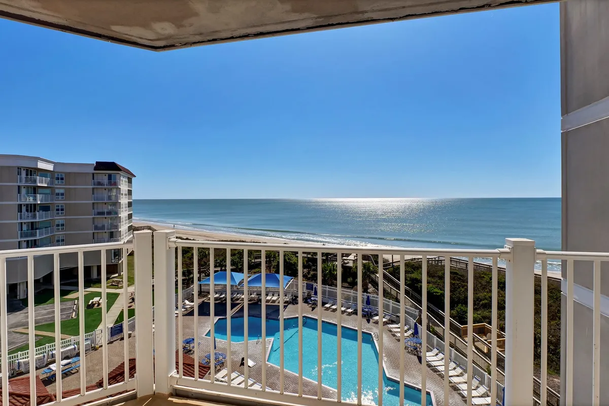 A scenic vista from a private balcony showing the sparkling pool area below, the expansive ocean beyond, and the inviting balcony railing in the foreground. The space is ideal for relaxing while overlooking both pool and beach.
