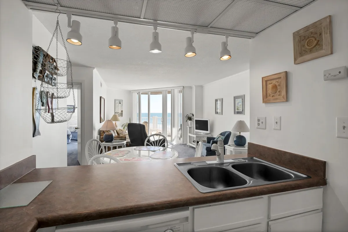 A bright, open kitchen area with a sink, a dining table, and windows framing a view of the ocean. Wall art adds a touch of personality, and the overall mood is both relaxed and inviting. The layout encourages cooking, dining, and casual conversation, while the ocean backdrop enhances the sense of being by the water.