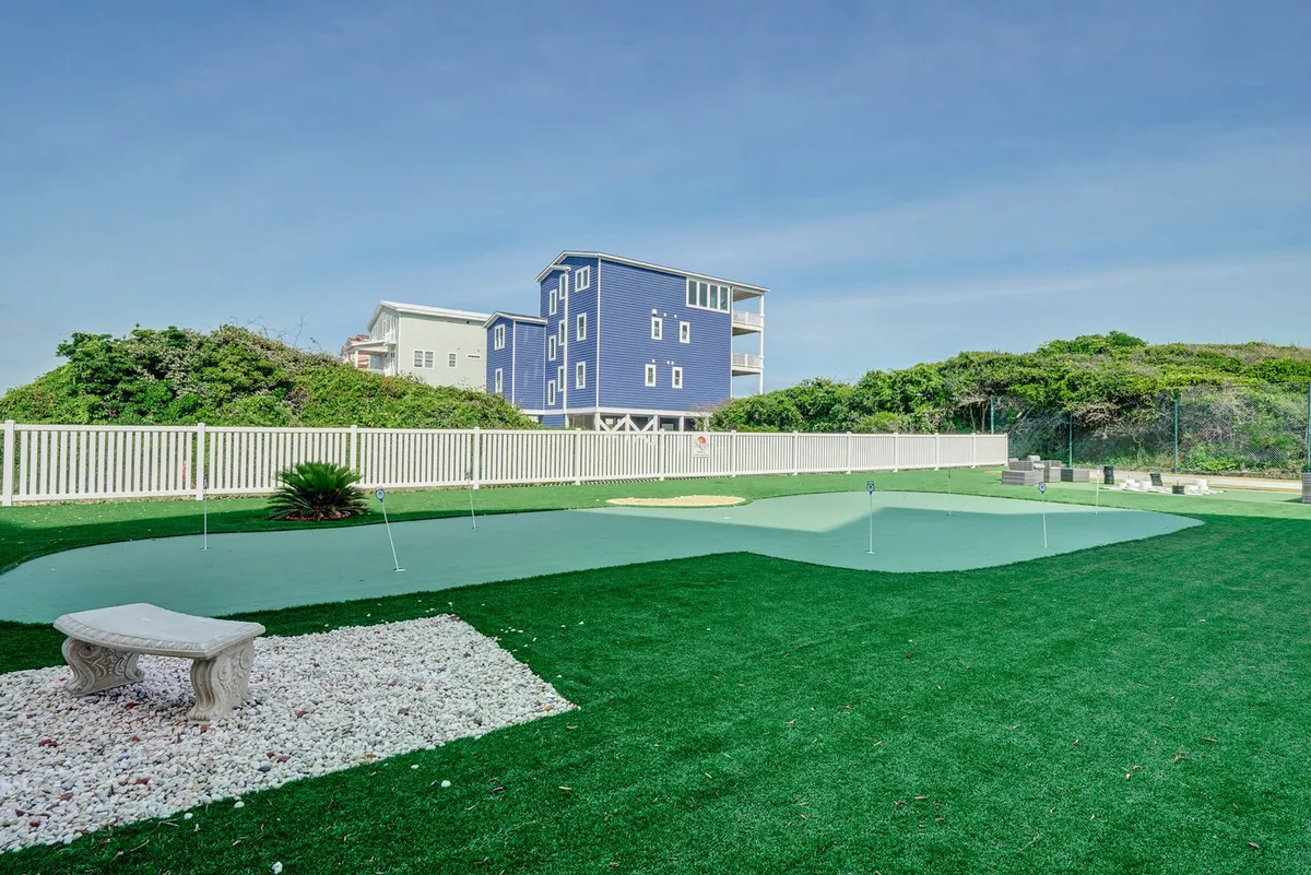 This image showcases a green putting green surrounded by white stone, lush plantings, and a blue house in the background. The space is perfect for practicing golf, socializing, or relaxing outdoors, encapsulating leisure activities for all ages.