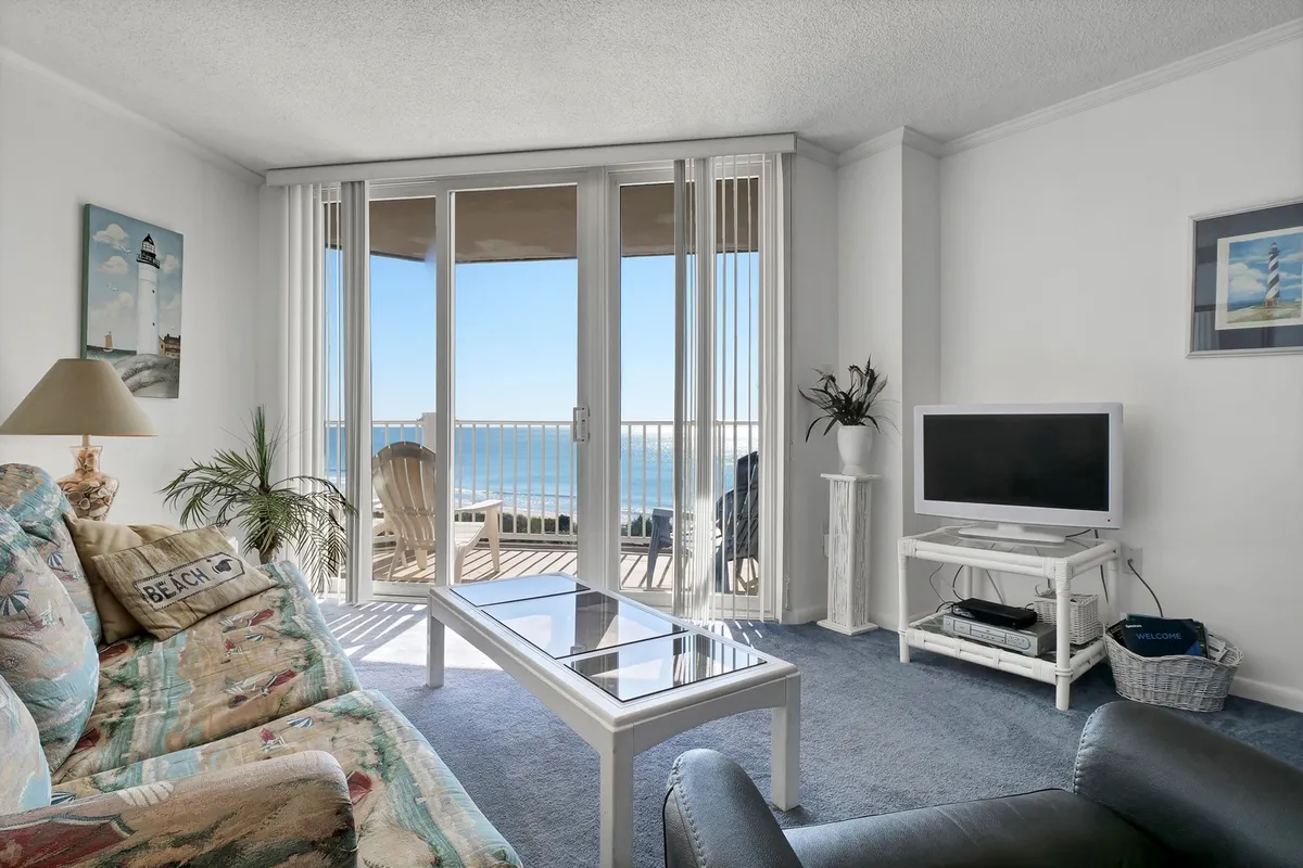 A spacious living room with large windows offering sweeping ocean views. The room features a comfortable couch, a coffee table, and a television, all arranged to invite relaxation and socializing. The windows let in abundant natural light, creating a tranquil and welcoming atmosphere. The balcony is visible through the glass, suggesting easy access to outdoor enjoyment.