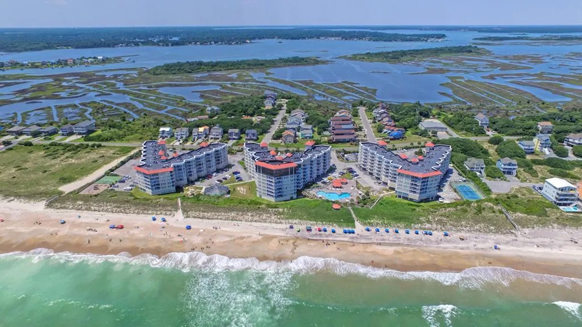 This exterior image captures a sweeping view of beachfront properties with a prominent swimming pool in the foreground, ocean waves rolling in the background, beach umbrellas set up for relaxation, and a marshland area nearby. The scene is vibrant, inviting, and clearly located at the edge of the natural environment.