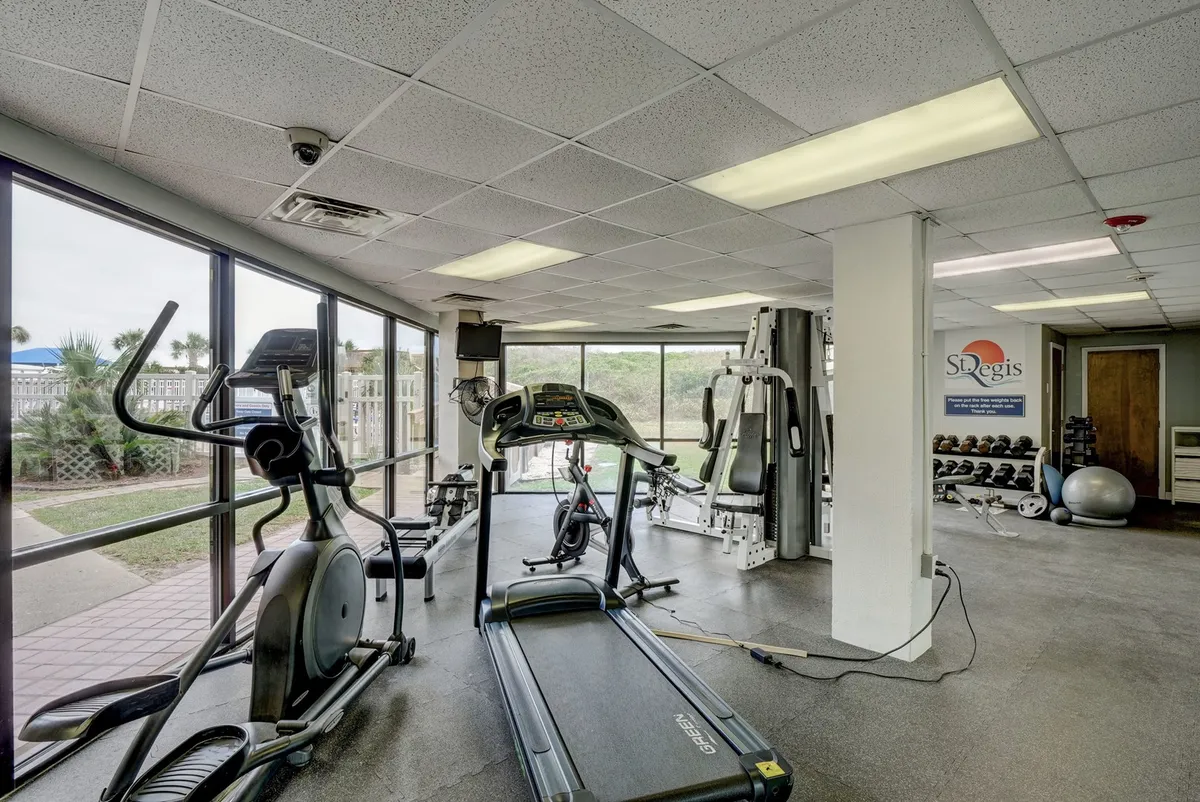 A bright, modern fitness center featuring a treadmill, elliptical machine, and weightlifting equipment set beside large windows that draw in plenty of natural light. The space is clean, motivating, and clearly designed for both cardio and strength workouts.