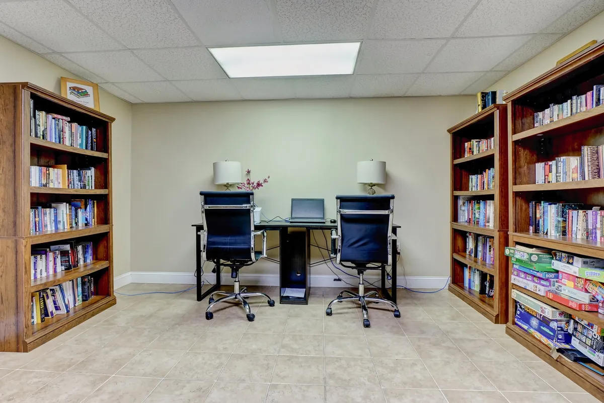 A serene business center with bookshelves brimming with books, two office chairs, and a small desk with a computer. The setting is calm, functional, and ideal for productivity or remote work.